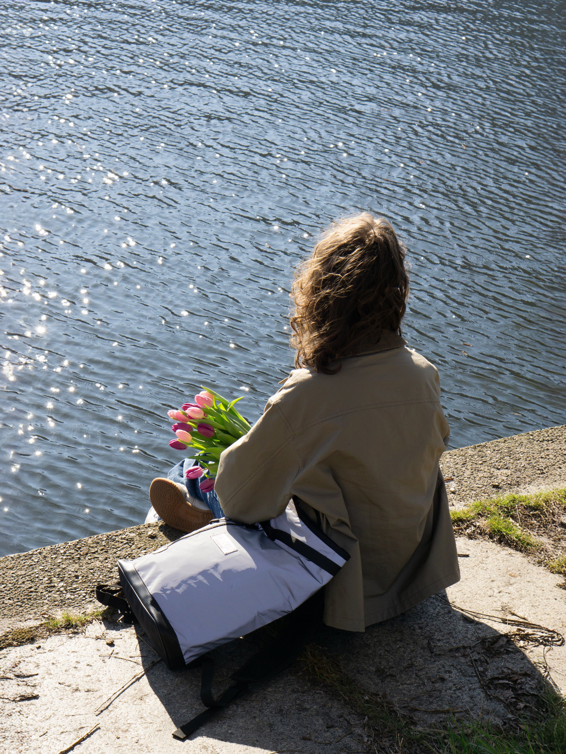 Eine Frau sitzt an der Spree mit dem Fahrradruckdsack Liz Small Glow – die reflektierende Fahrradtasche und Rucksack in einem für stilbewusste Radfahrer:innen.