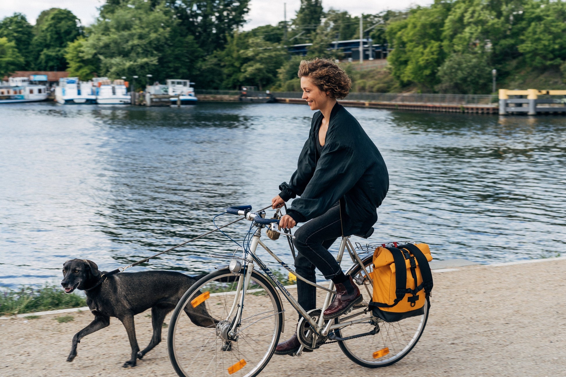 Frau fährt Fahrrad an der Spree, trägt Rucksack seitlich als Fahrradtasche sicherer befestigt mit der Fahrradhalterung. Sie hält einen Hund an der Leine befestigt. Urbaner Lifestyle, praktische Fahrradlösung. ©Spleen Berlin